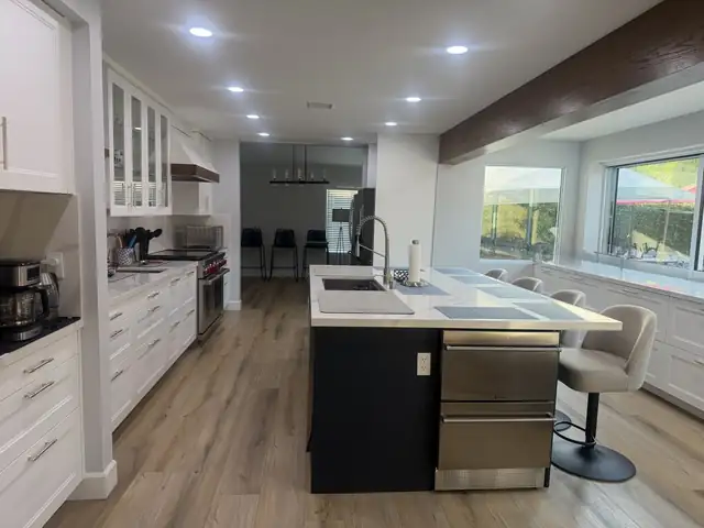 Open white kitchen with a navy island, stainless dishwasher, wood ceiling beam and wood-look floor