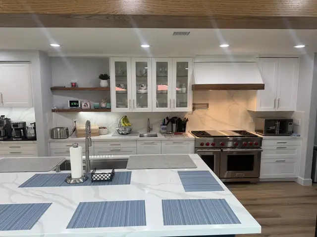 White shaker kitchen with glass-front upper cabinets, open wood shelves, a white quartz island and a range hood with a wood band